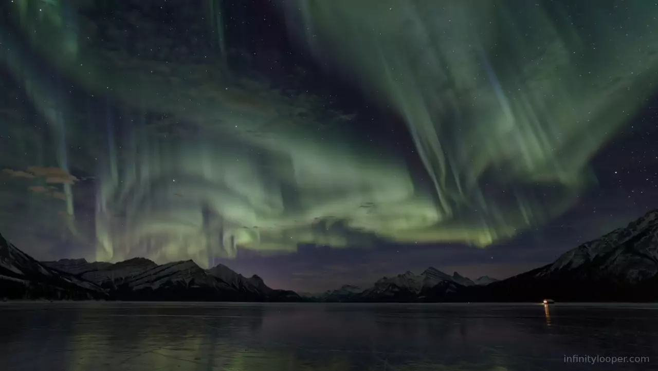Green aurora across a starry sky above dark snowy mountains and a still icy lake with a small light on the shore